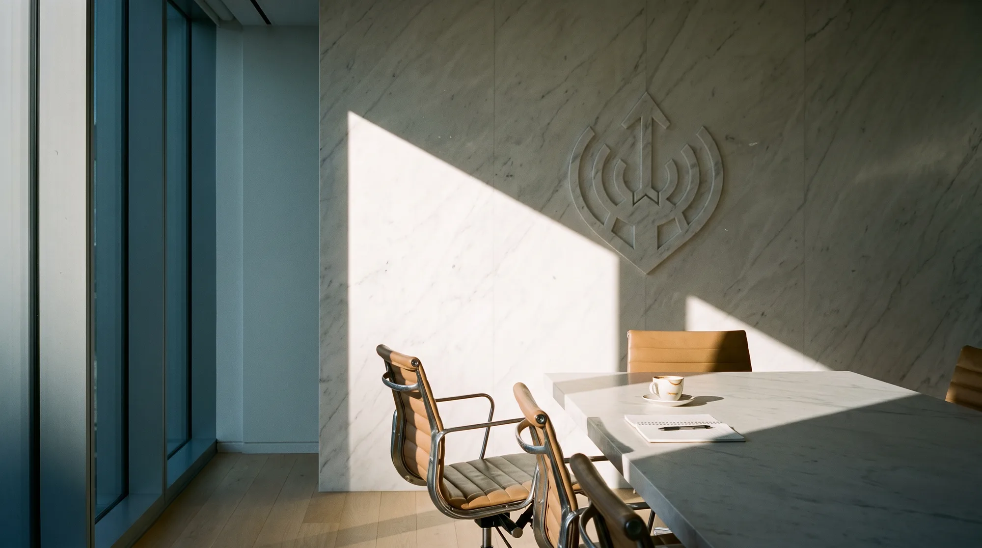 Late afternoon light across the MANTICO Group boardroom, the emblem engraved into the marble wall